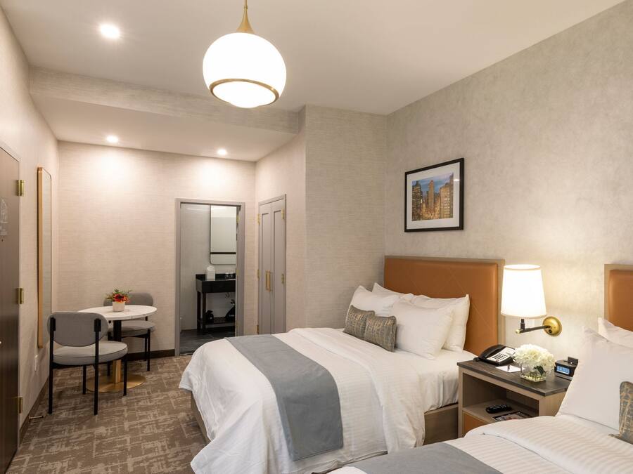 A hotel room with two beds, a desk with a chair, a small table with flowers, a lamp, and a framed picture on the wall.