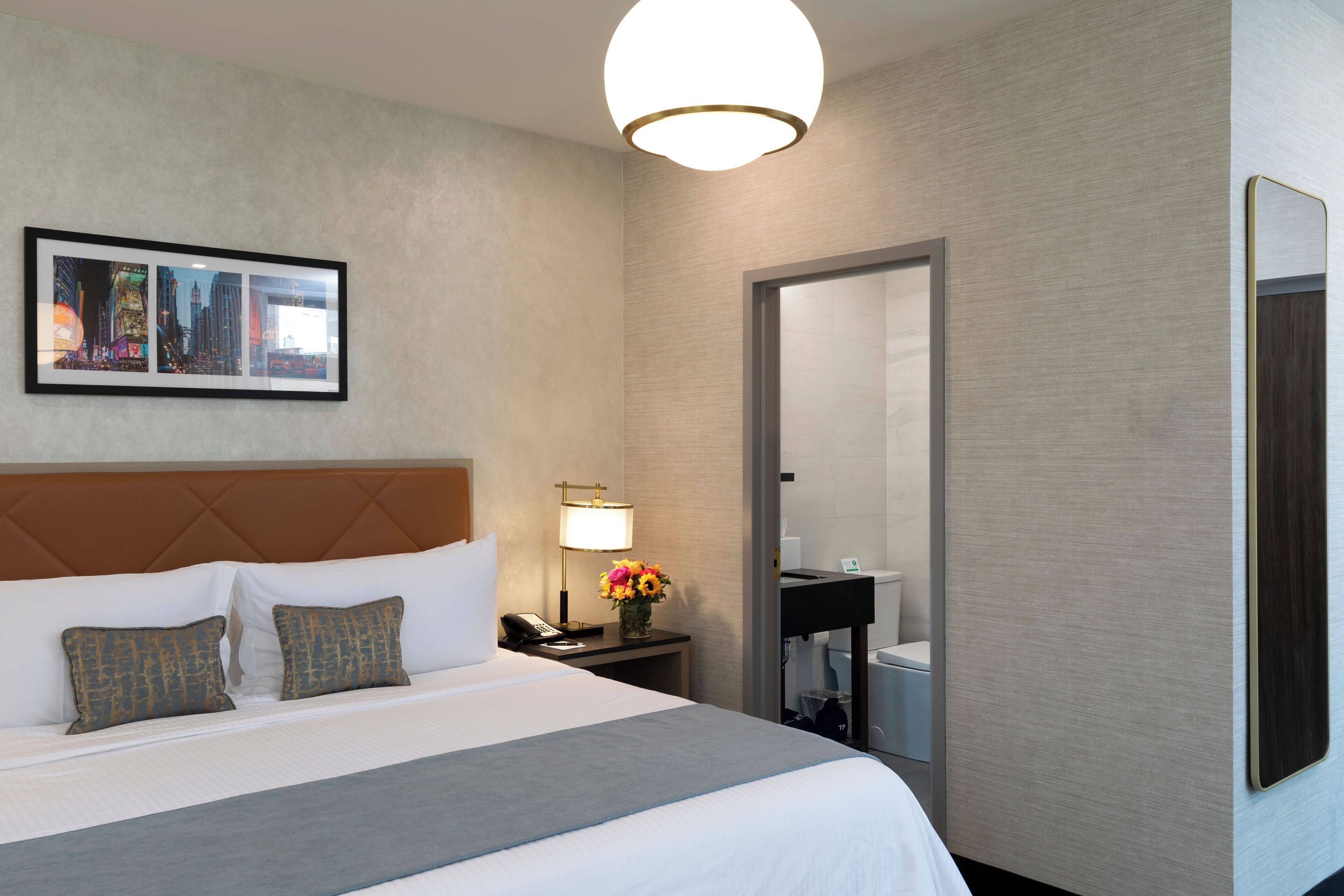 A hotel room with a large bed, a nightstand with a lamp, a framed picture on the wall, and a bathroom visible through an open door.