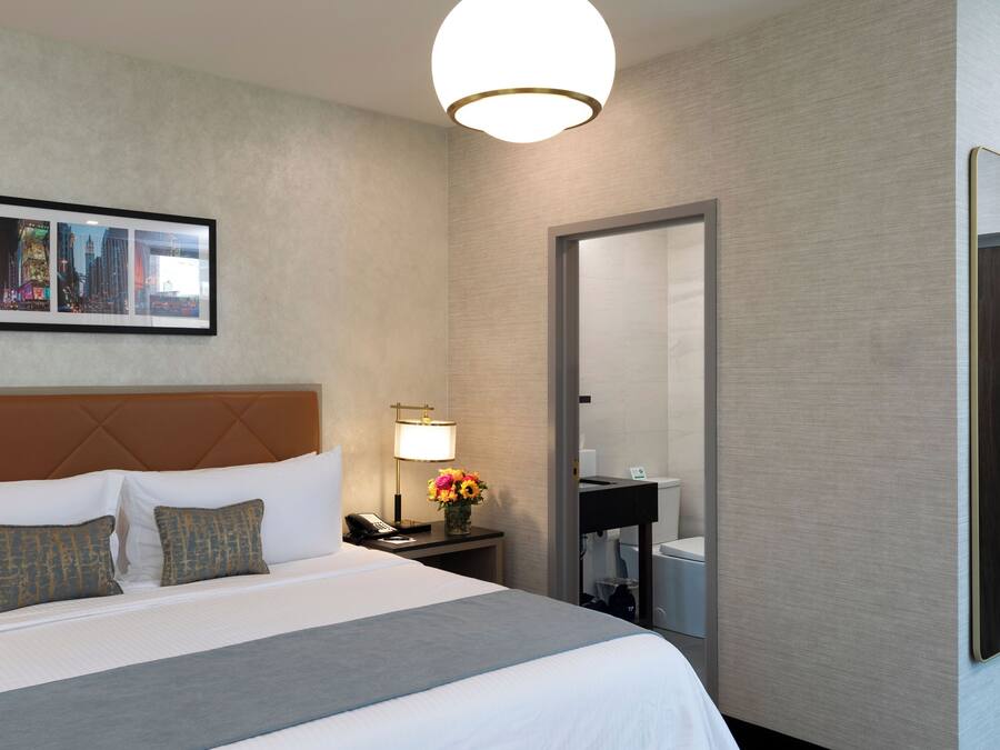 Habitación de hotel con una cama grande, una mesita de noche con lámpara, un cuadro en la pared y un baño visible a través de una puerta abierta.