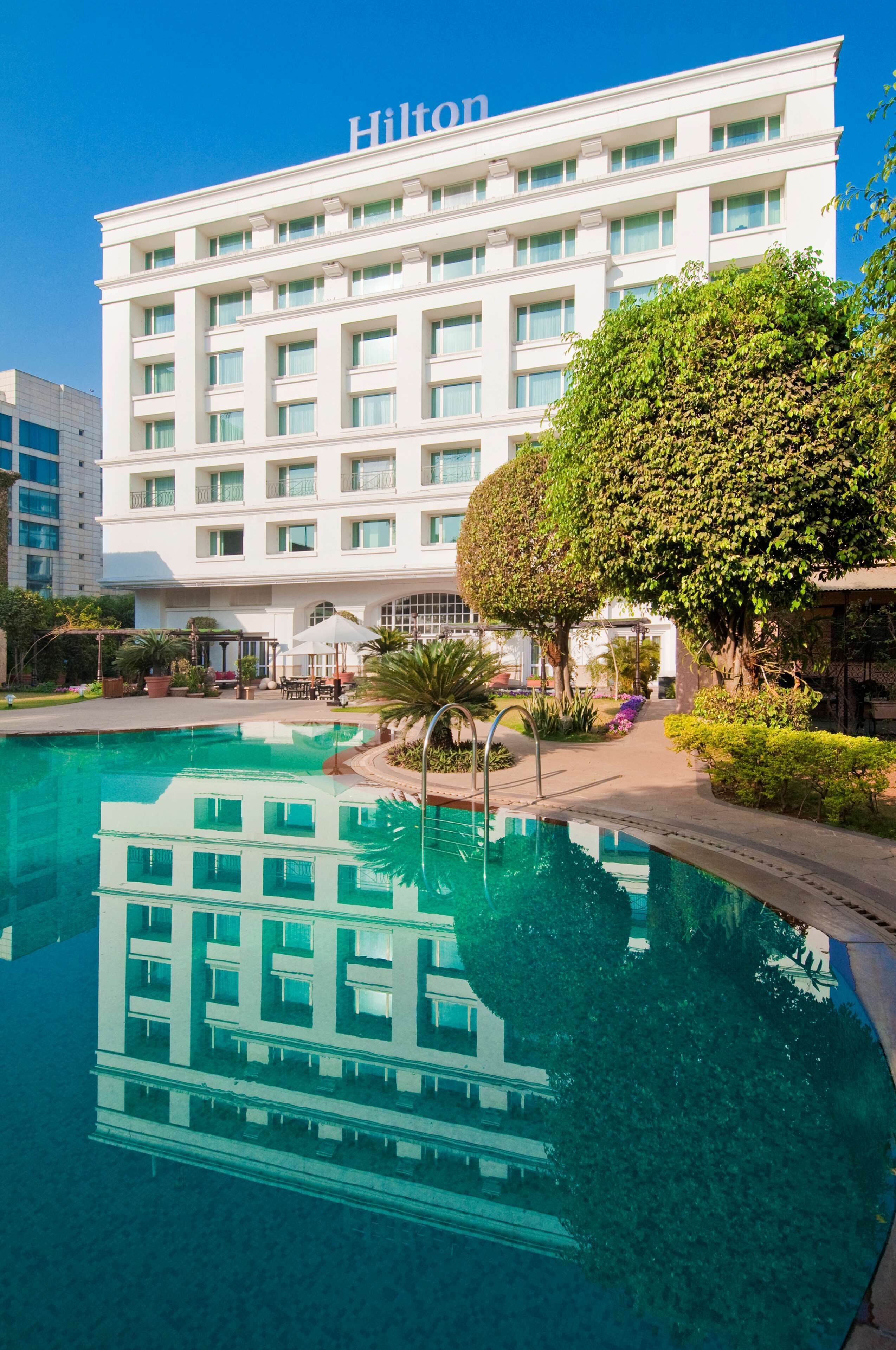 A hotel with a swimming pool, surrounded by greenery and a clear blue sky.