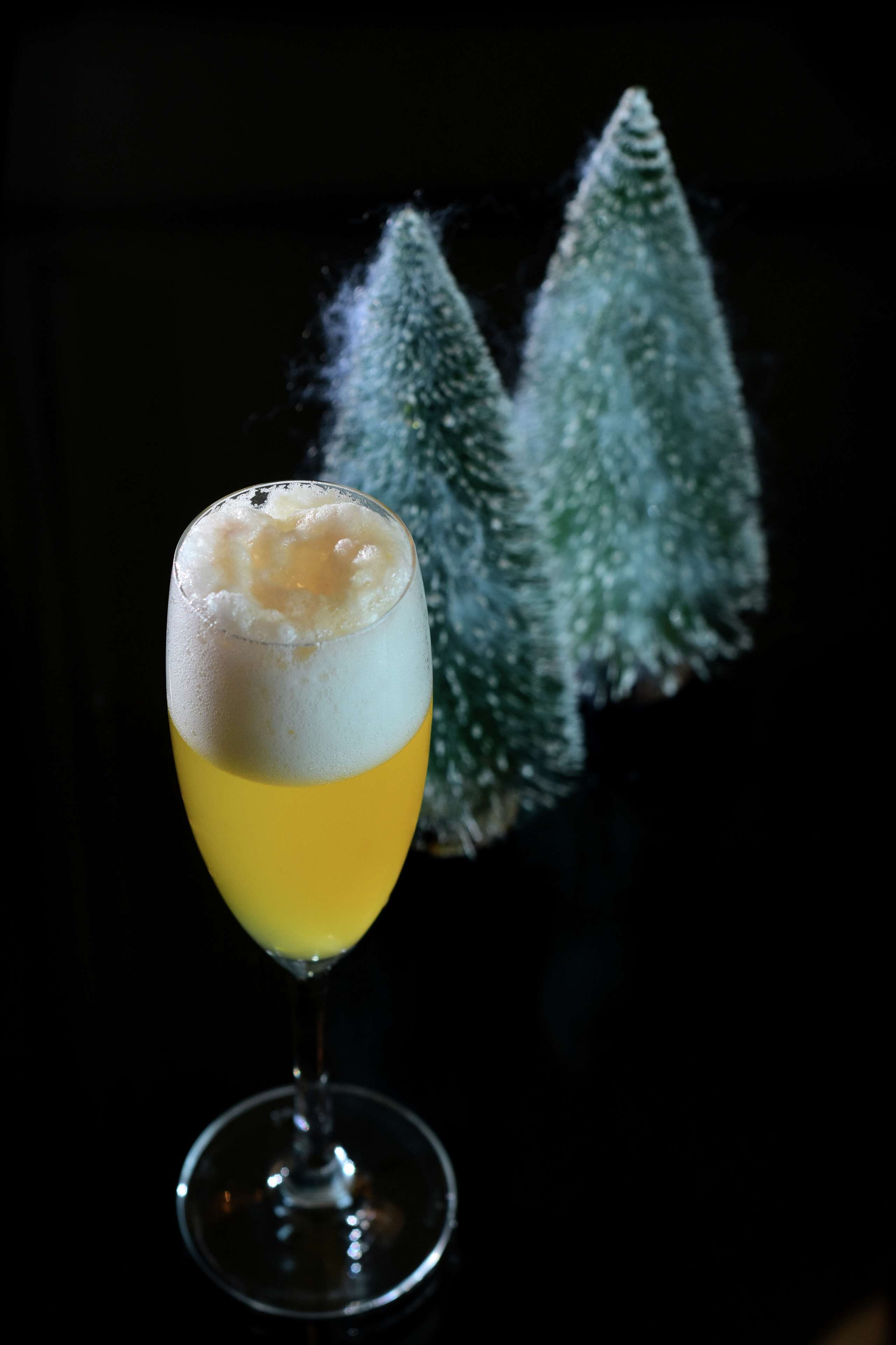 A cocktail in a stemmed glass with a frothy top, set against a dark background with two conical decorations.