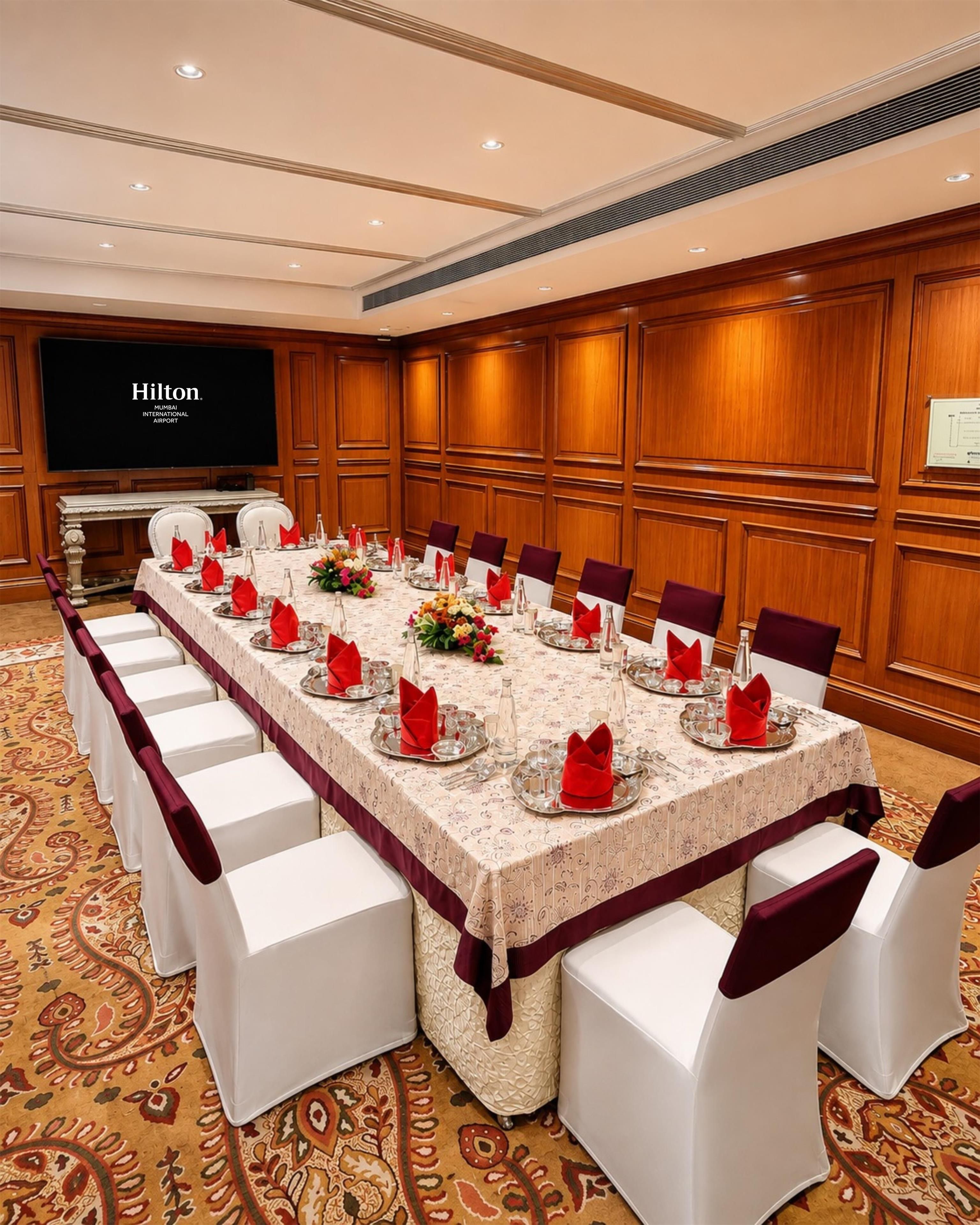 A conference room with a long table set for a meeting, white chairs, and a large screen displaying 'Hilton Mumbai International Airport'.