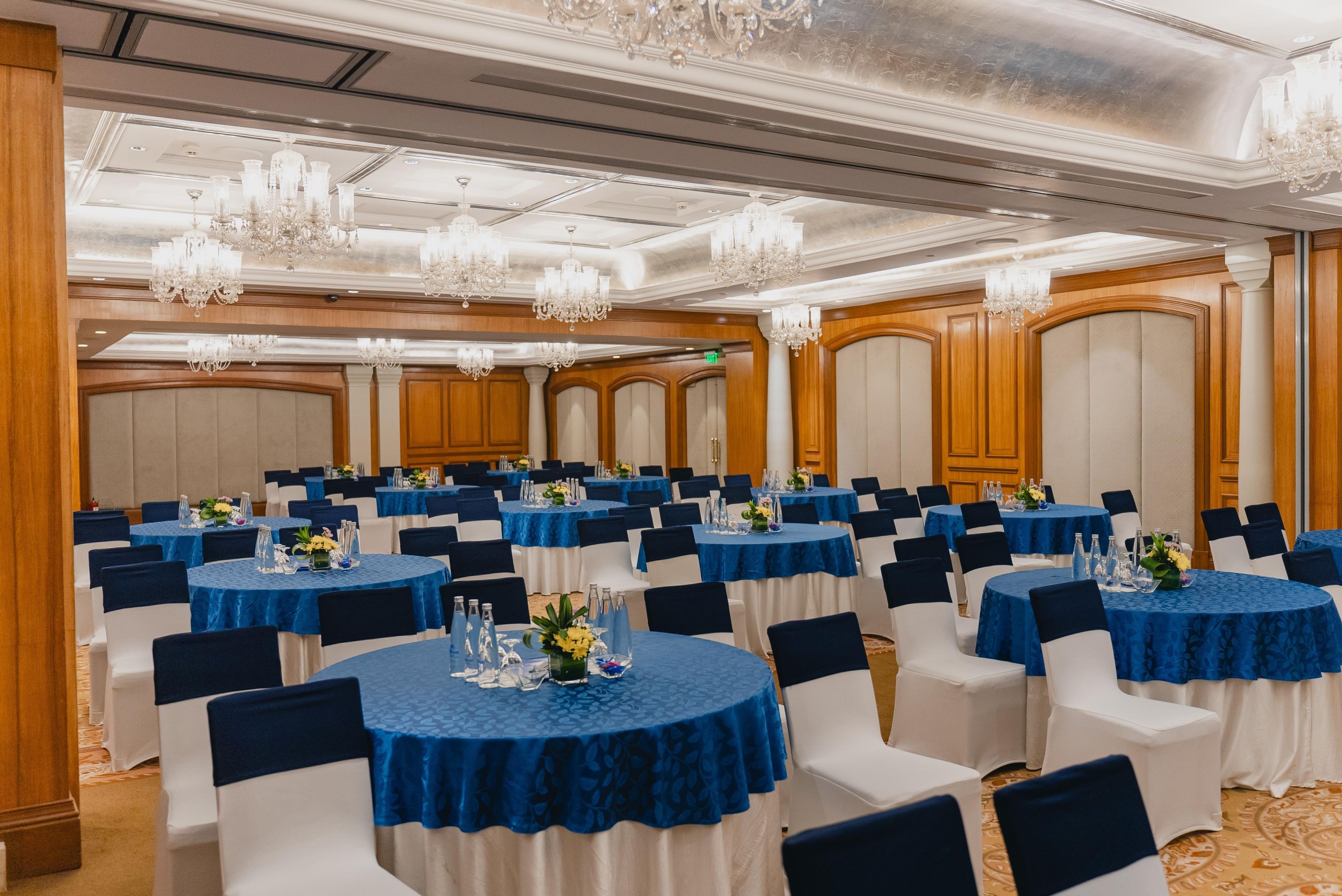 A banquet hall with round tables, blue tablecloths, white chairs, and chandeliers.