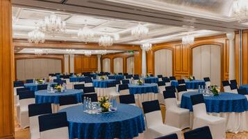 A banquet hall with round tables, blue tablecloths, white chairs, and chandeliers.