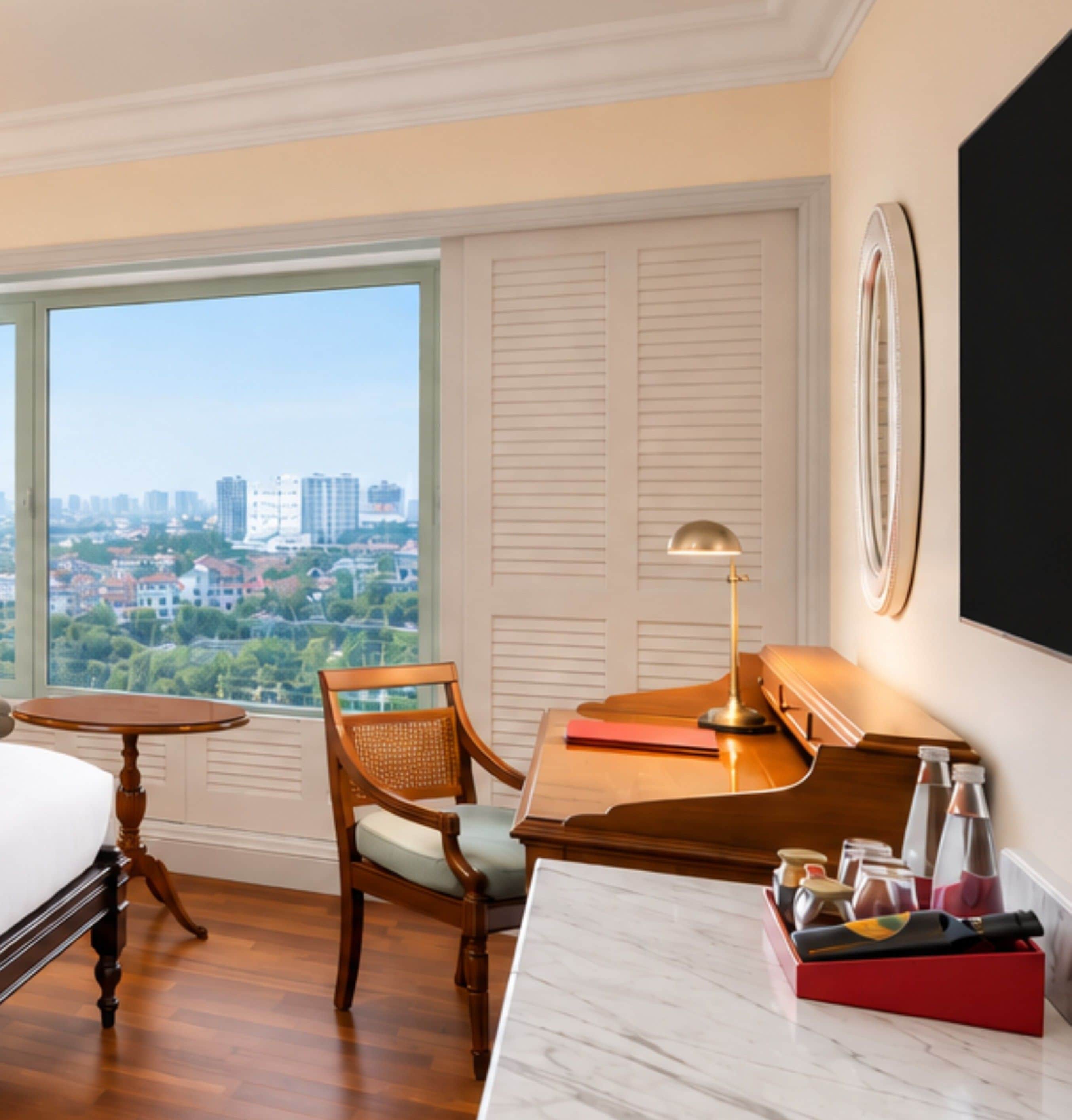 A hotel room with a large window, a desk with a lamp, a chair, a bed, and a view of a cityscape.
