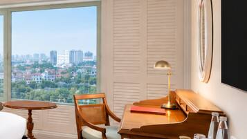 A hotel room with a large window, a desk with a lamp, a chair, a bed, and a view of a cityscape.