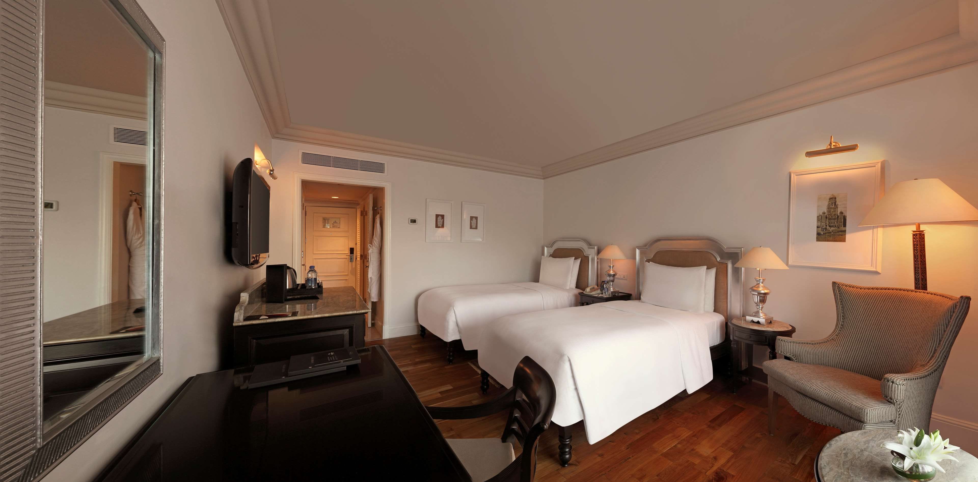 A hotel room with two beds, a desk with a flat-screen TV, a chair, and a small table with a vase of flowers.