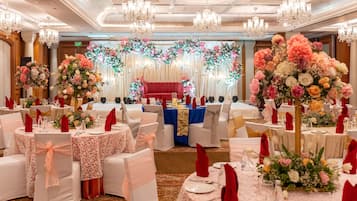Un salón de banquetes con mesas redondas, sillas blancas adornadas con cintas rosas y centros de mesa florales.