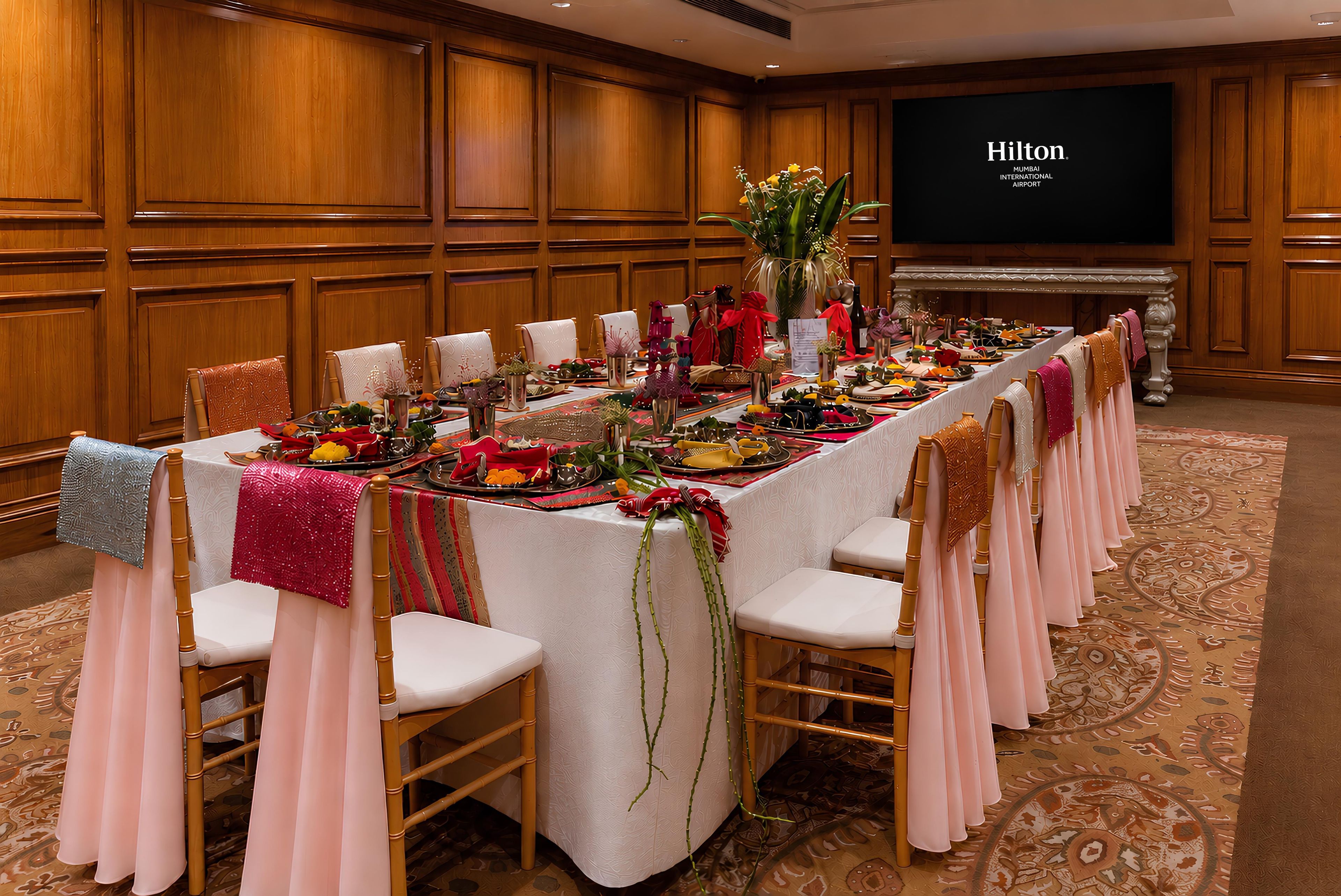 A long banquet table set for a formal event, with chairs on both sides, in a room with wooden paneling and a large screen displaying the Hilton logo.