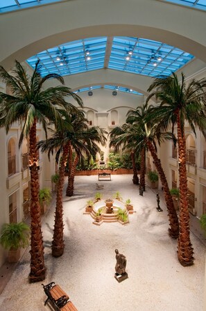 Un vaste atrium doté d’un haut plafond, agrémenté de palmiers, d’une fontaine centrale et d’une statue.
