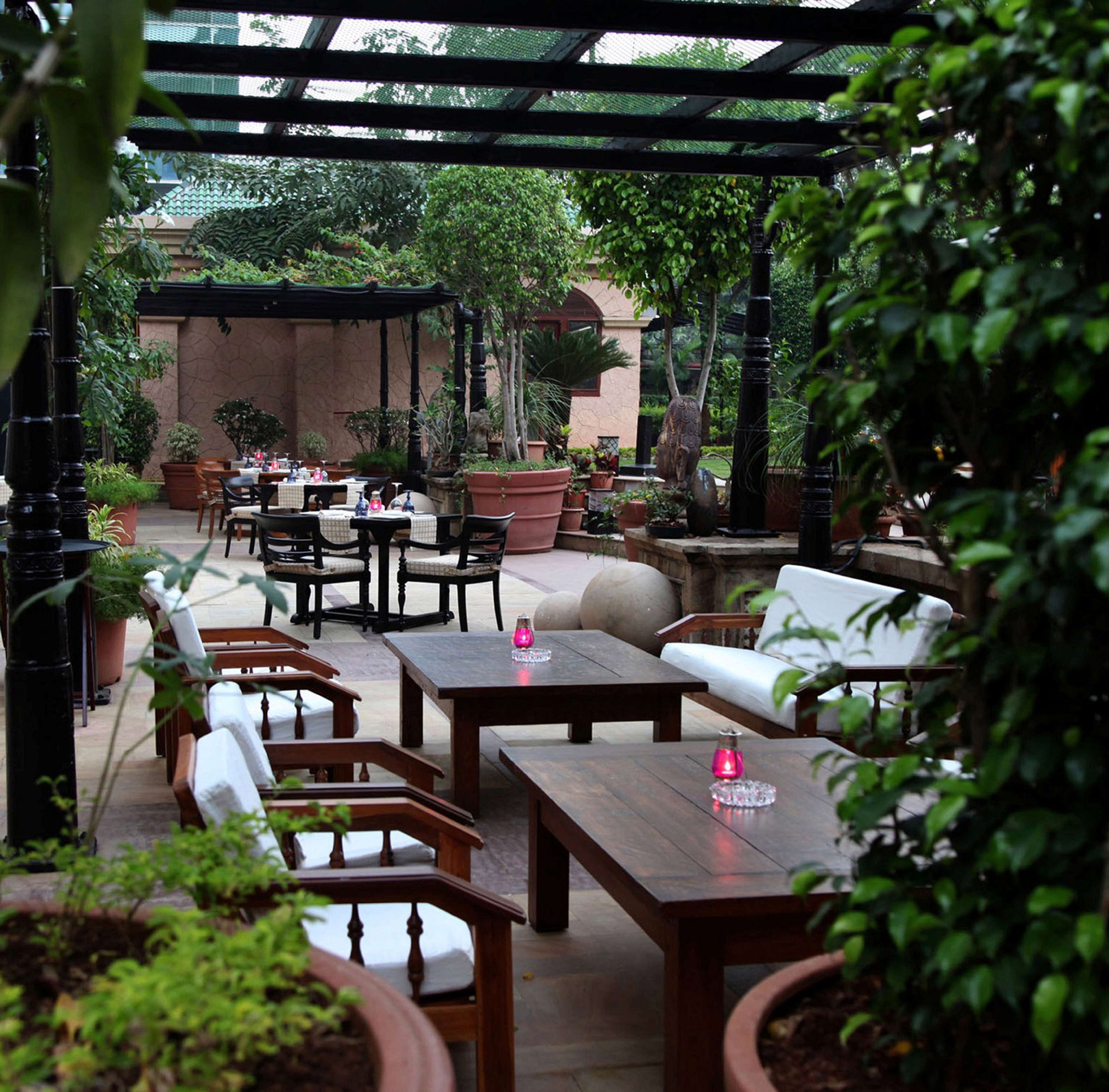 An outdoor patio area with wooden furniture, including sofas and tables, surrounded by potted plants and a pergola structure.