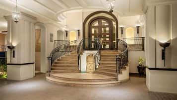 Un amplio vestíbulo de hotel con una escalera central, lámparas ornamentales y un gran arco de entrada.