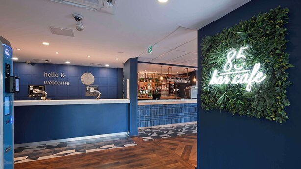 A reception area with a blue wall, a clock, and a neon sign reading "85 bar cafe". - Travelodge Bracknell Central (Bracknell)