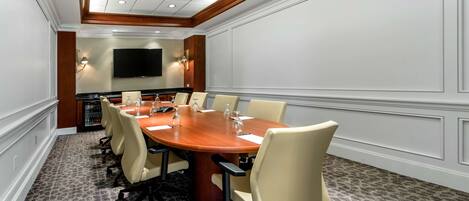 A conference room with a large wooden table, beige chairs, a flat-screen TV mounted on the wall, and a minibar.