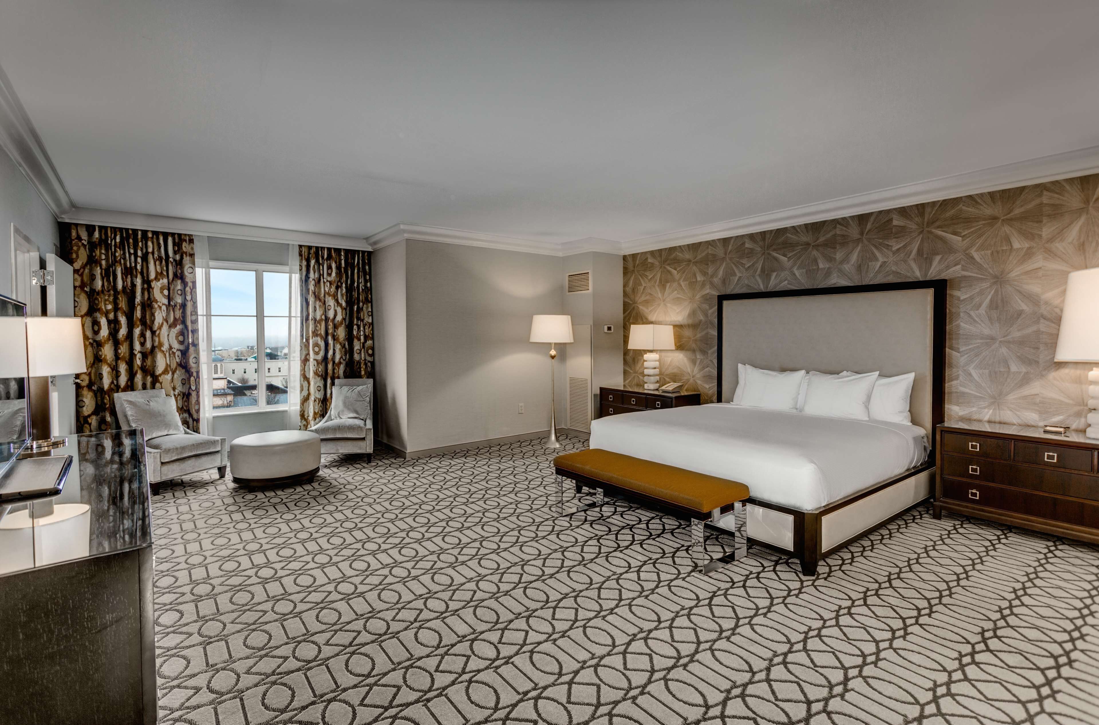 A hotel room with a large bed, a desk with a computer, a chair, a bench, and a view of the city through large windows.