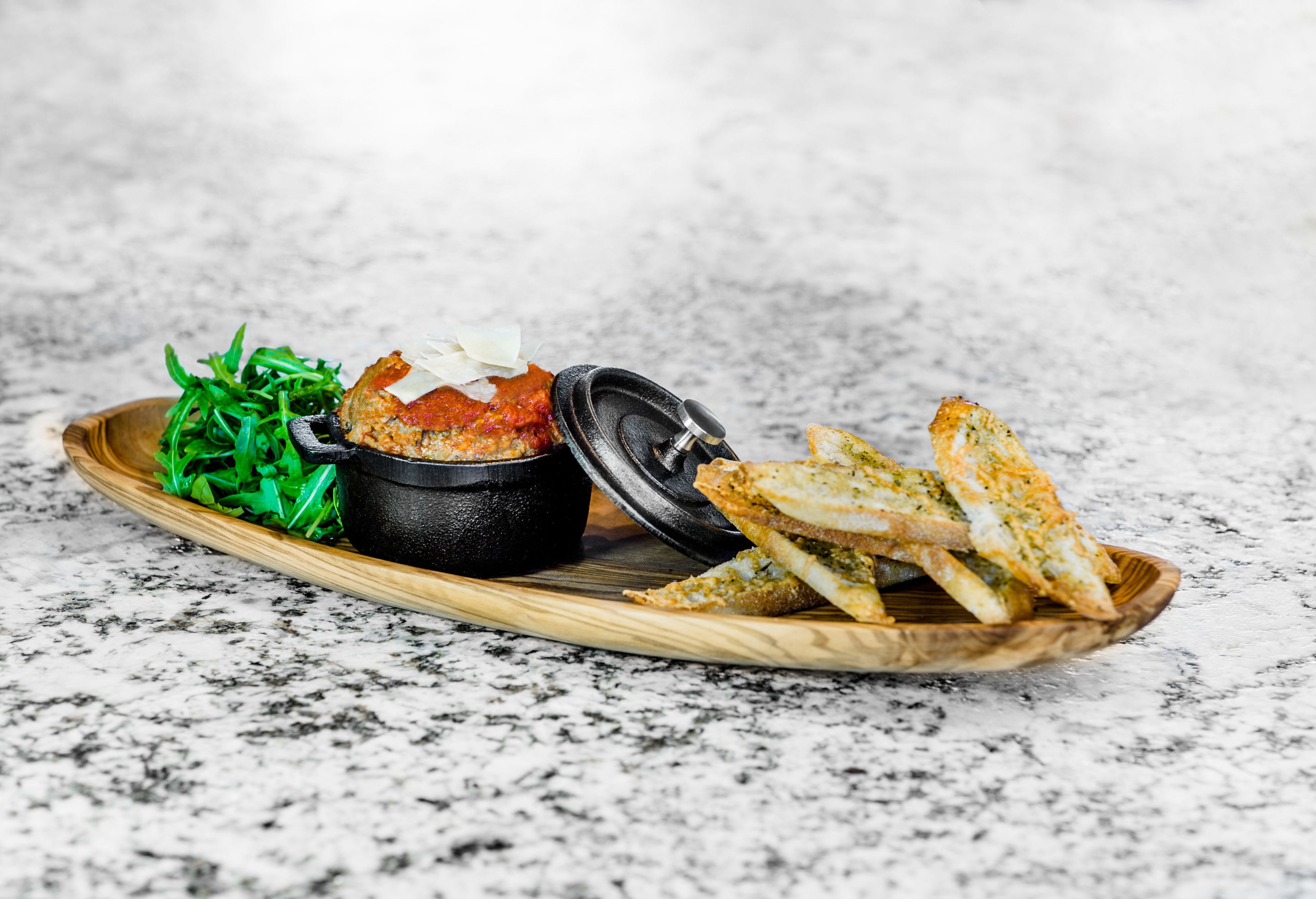 A wooden tray with a black bowl, a piece of bread, and green salad on a marble surface.