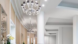 A spacious hotel corridor with a marble floor, large chandeliers, and a glass railing overlooking a balcony.