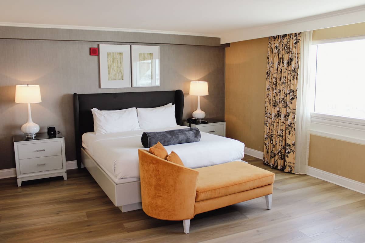 Una habitación de hotel con una cama, dos mesitas de noche con lámparas, un aparador, un ventanal con cortinas y suelo de madera.