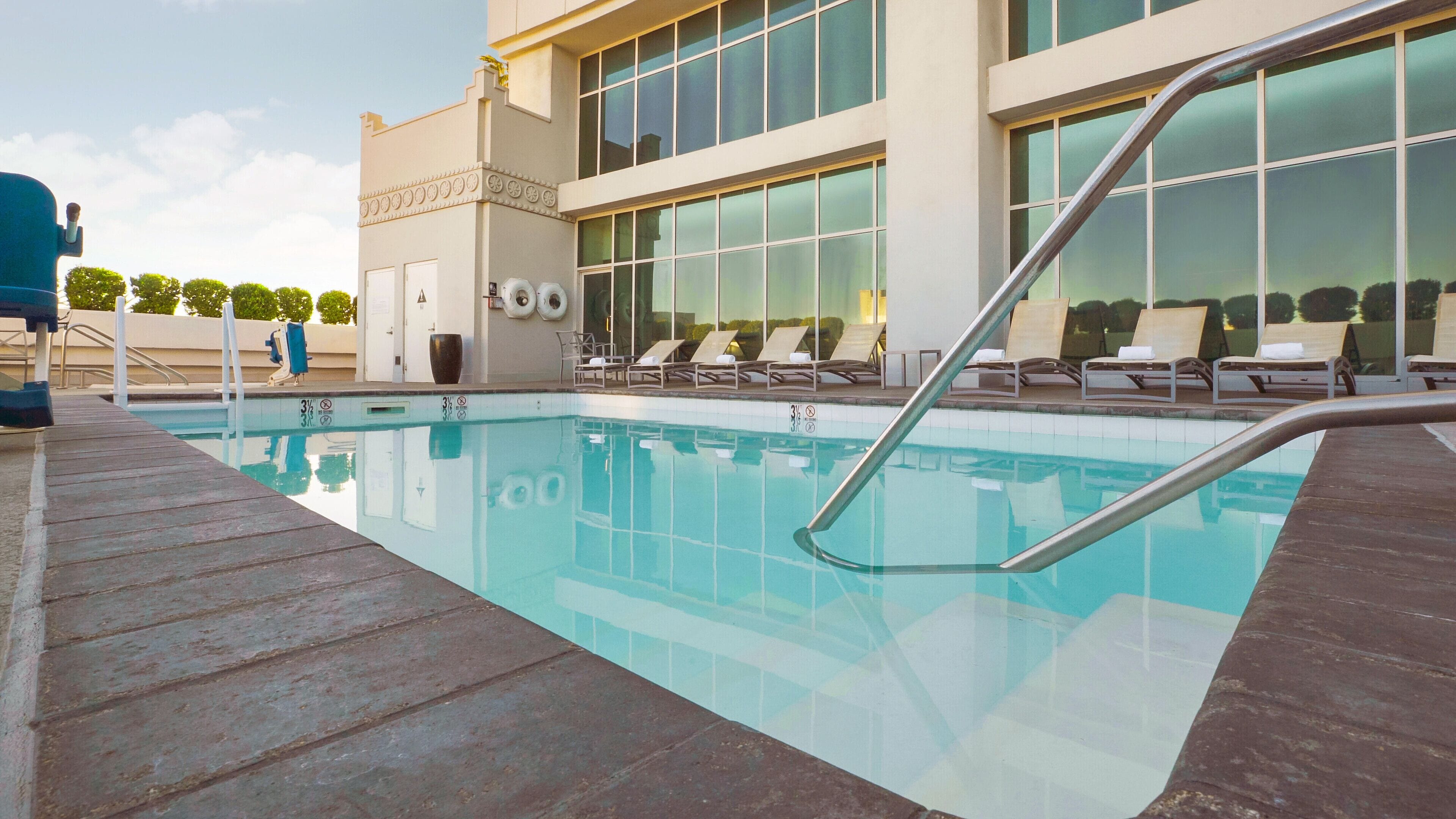 Una piscina con cubierta de madera, sillas de descanso y un edificio con ventanas amplias al fondo.