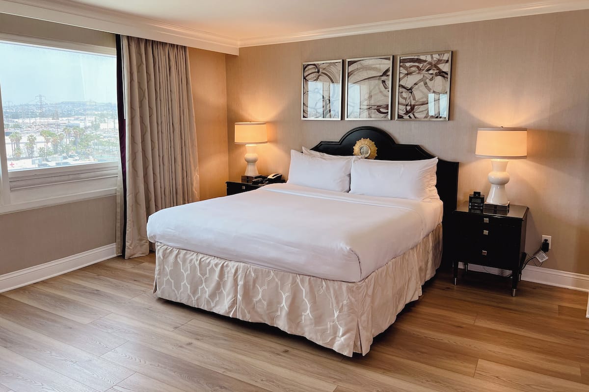 Habitación de hotel con una cama grande, dos mesitas de noche con lámparas, vista a la ciudad por una ventana y tres obras de arte colgadas en la pared.