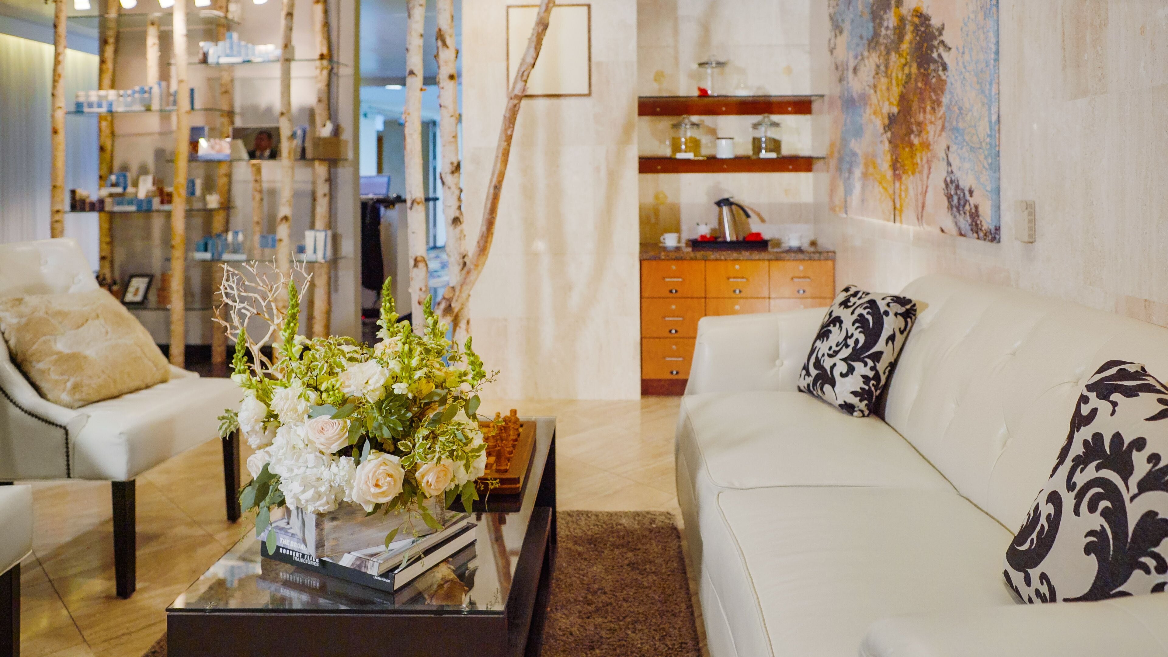 Un salón moderno con un sofá blanco, una mesa baja con un arreglo floral y una estantería al fondo.