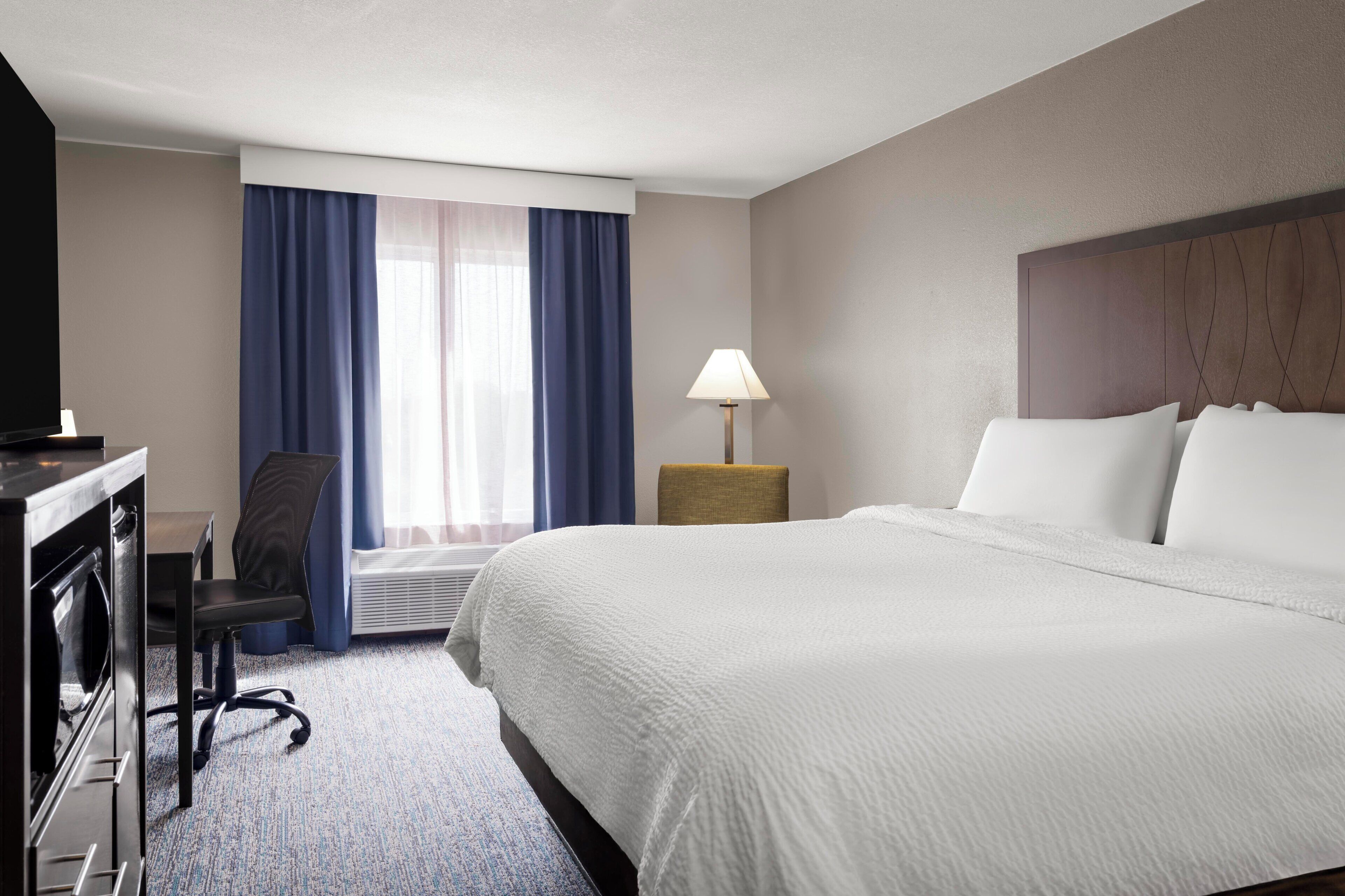 A hotel room with a bed, a desk, a chair, a lamp, and a window with curtains.