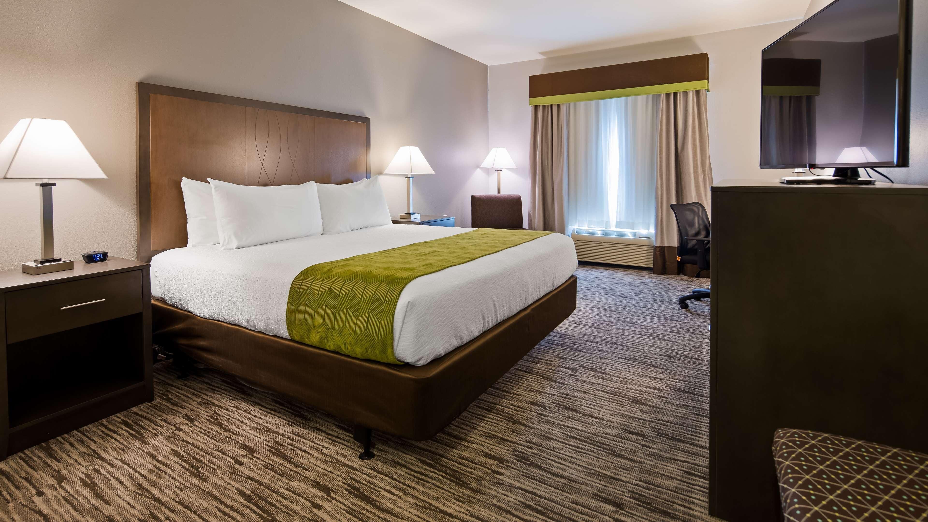 A hotel room with a large bed, two bedside lamps, a desk with a lamp, a chair, a television, and a window with curtains.