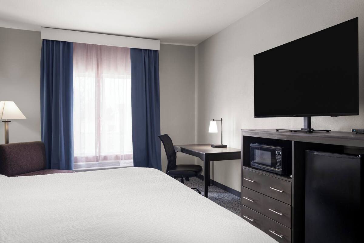 A hotel room with a bed, a desk with a chair, a TV, a microwave, and a refrigerator.