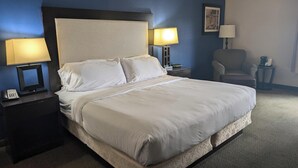 Una cama bien hecha con sábanas blancas en una habitación con paredes azules. Hay dos mesitas de noche con lámparas, una silla y una pequeña mesa con un teléfono y una cafetera.