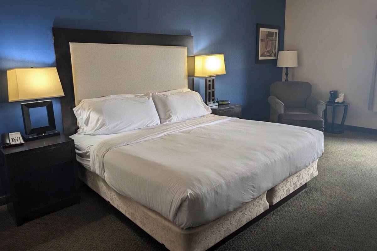 Una cama bien hecha con sábanas blancas en una habitación con paredes azules. Hay dos mesitas de noche con lámparas, una silla y una pequeña mesa con un teléfono y una cafetera.