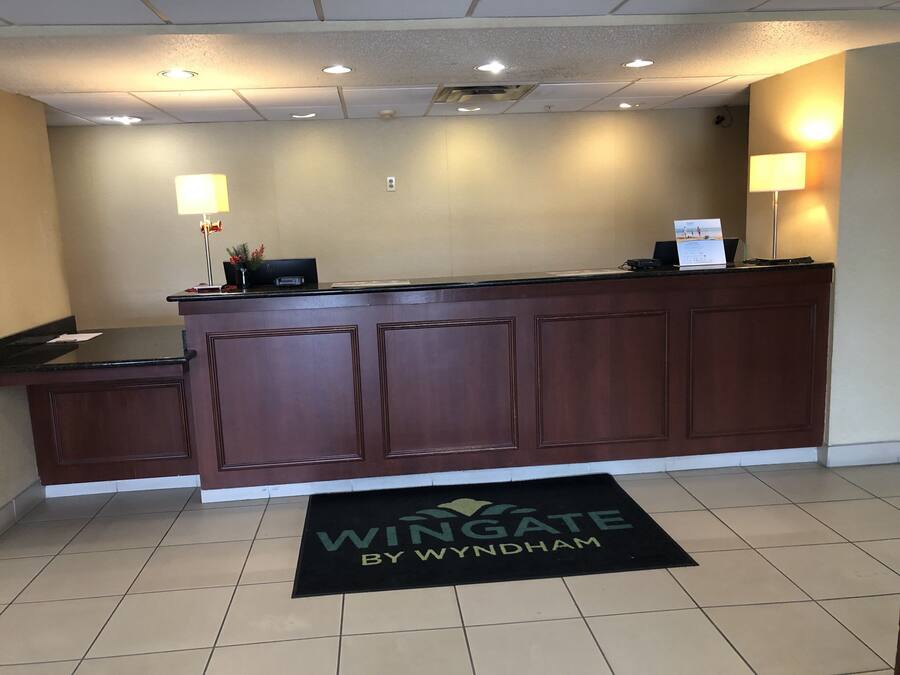 Un mostrador de recepción con acabado de madera oscura, un tapete negro con el logotipo de 'Wingate by Wyndham' y dos lámparas de pared.