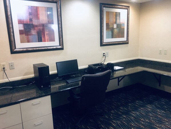 A small office area with a desk, computer, and printer. There are framed artworks on the wall and a carpeted floor.