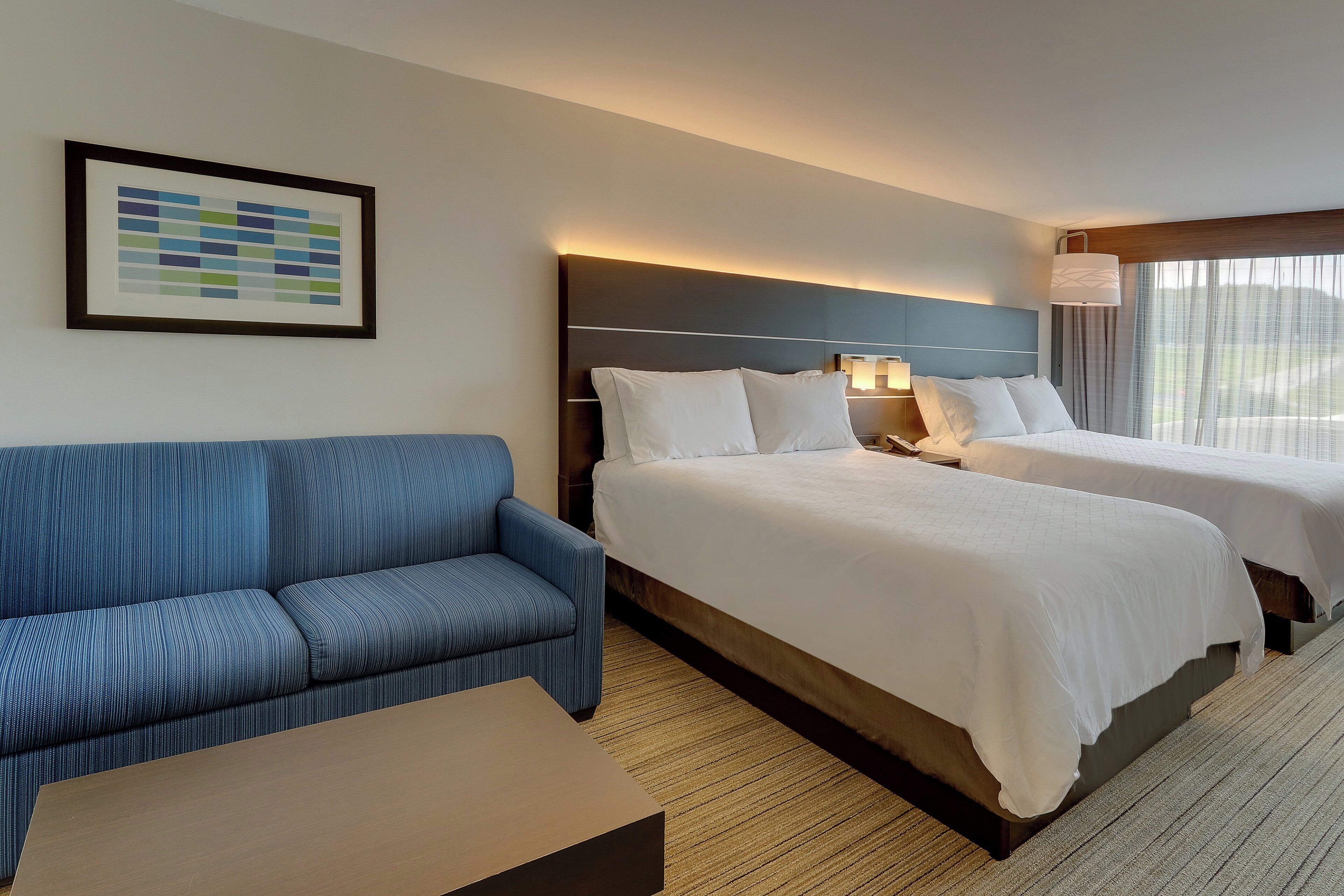 Habitación de hotel con sofá azul, dos camas, un cuadro en la pared y una ventana con cortinas.