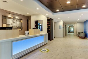 Uma área de recepção de hotel com design moderno, balcão de atendimento, tela digital e corredor que dá acesso a outras áreas.