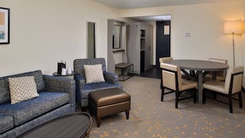 Habitación de hotel con sofá, reposapiés, mesa redonda y sillas. Hay una mesita de noche con una lámpara y un cuadro en la pared.