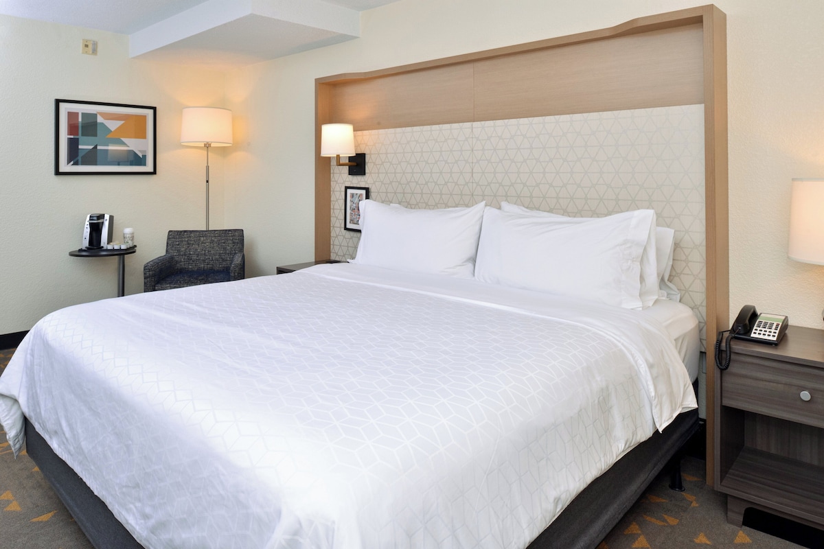 Una habitación de hotel con una cama grande, una silla, una mesita de noche con teléfono y una lámpara de pared.