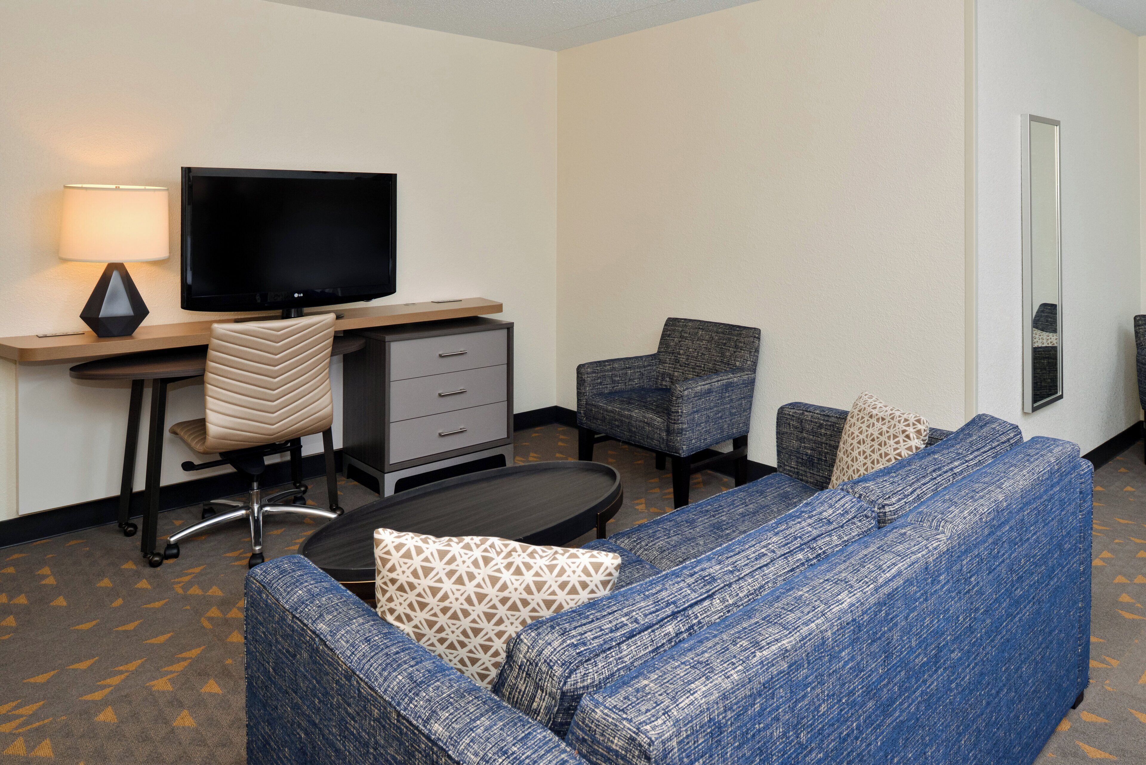 A hotel room with a large flat-screen TV, a desk with a lamp, a sofa, two chairs, and a small table.