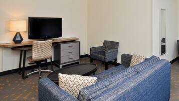 A hotel room with a large flat-screen TV, a desk with a lamp, a sofa, two chairs, and a small table.