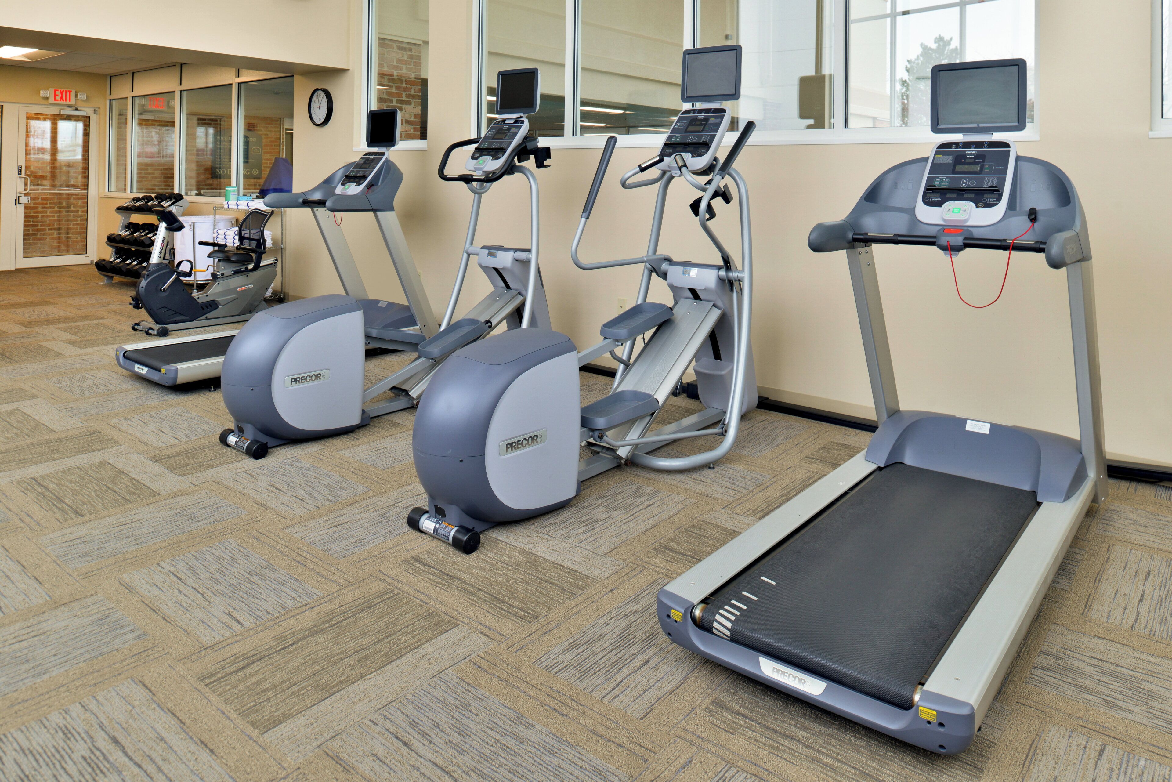 Eine Reihe von Laufbändern und Crosstrainern in einem Fitnessbereich mit Teppichboden und einem Fenster mit Jalousien.