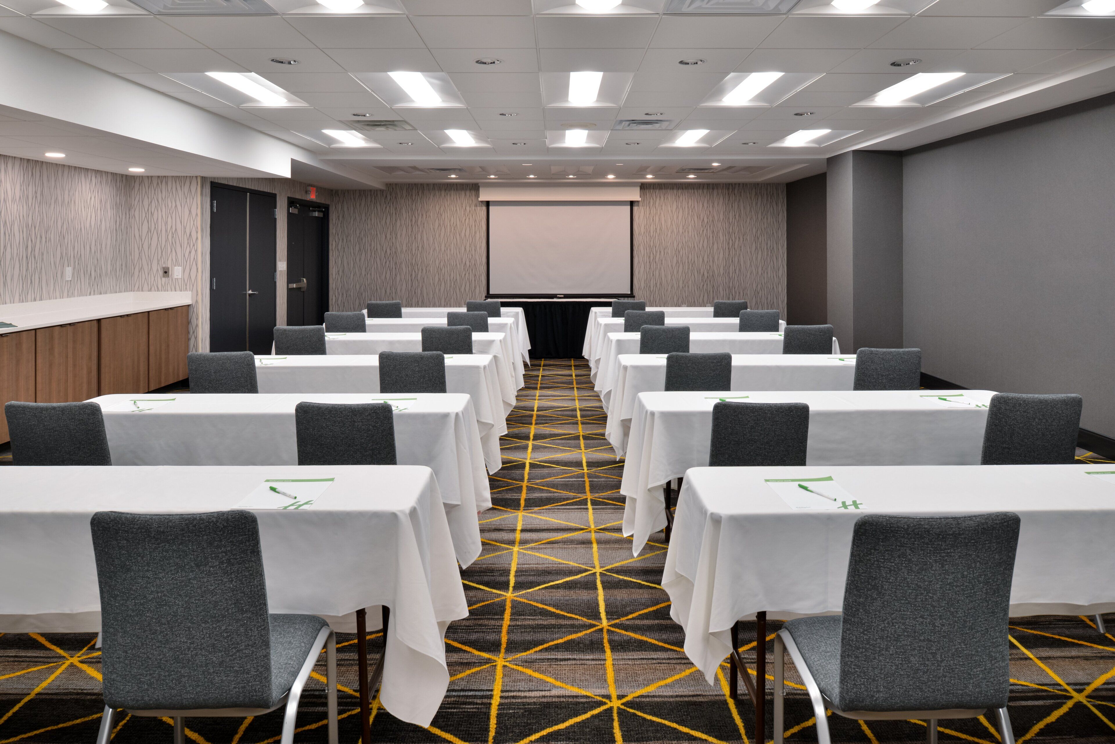 Une salle de conférence avec des rangées de tables et de chaises, un écran de projection et un sol recouvert de moquette.