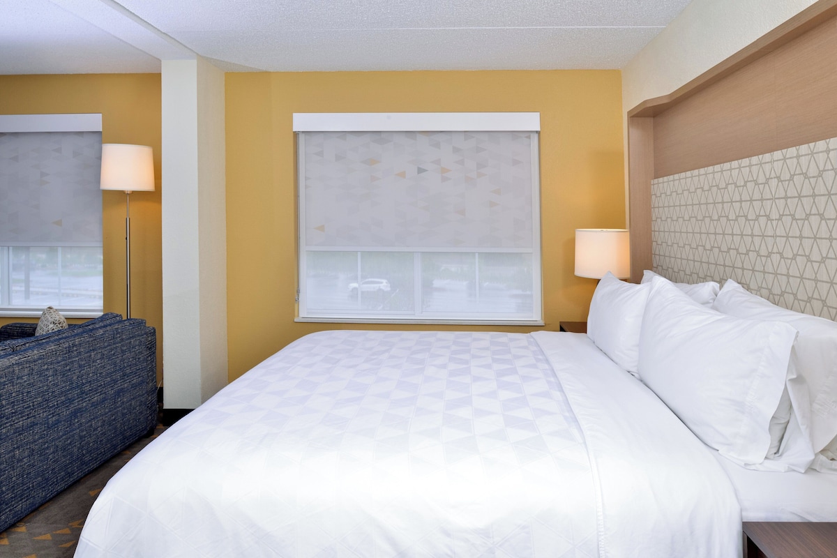 Una habitación de hotel con una cama grande, un sofá, una ventana con persianas y una lámpara.