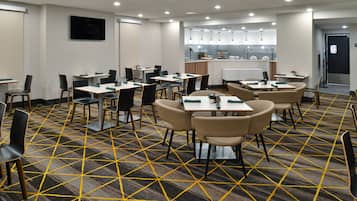 A conference room with tables and chairs, a kitchen area, and a patterned carpet.
