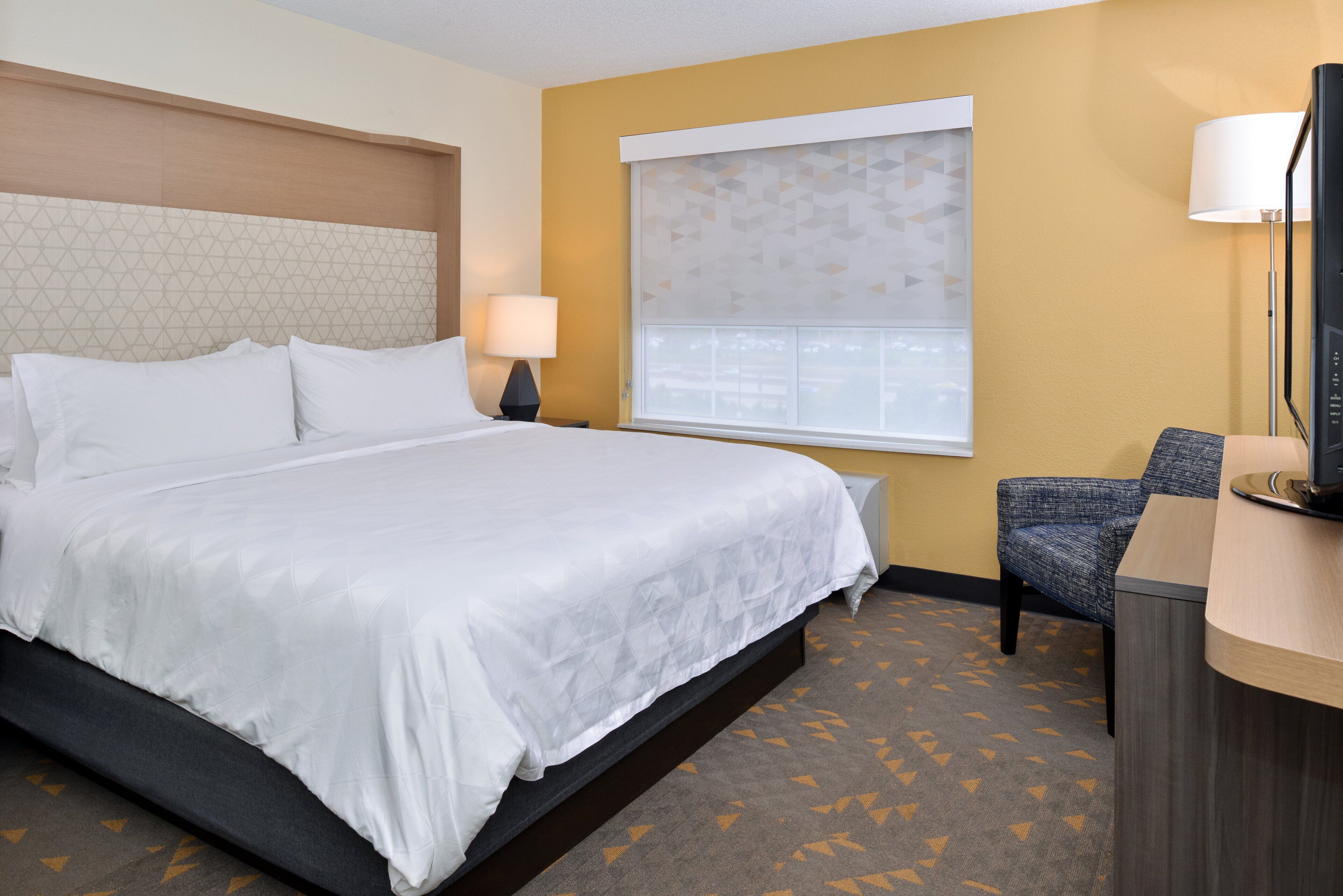 A hotel room with a large bed, a desk with a chair, a window with blinds, and a television on the desk.
