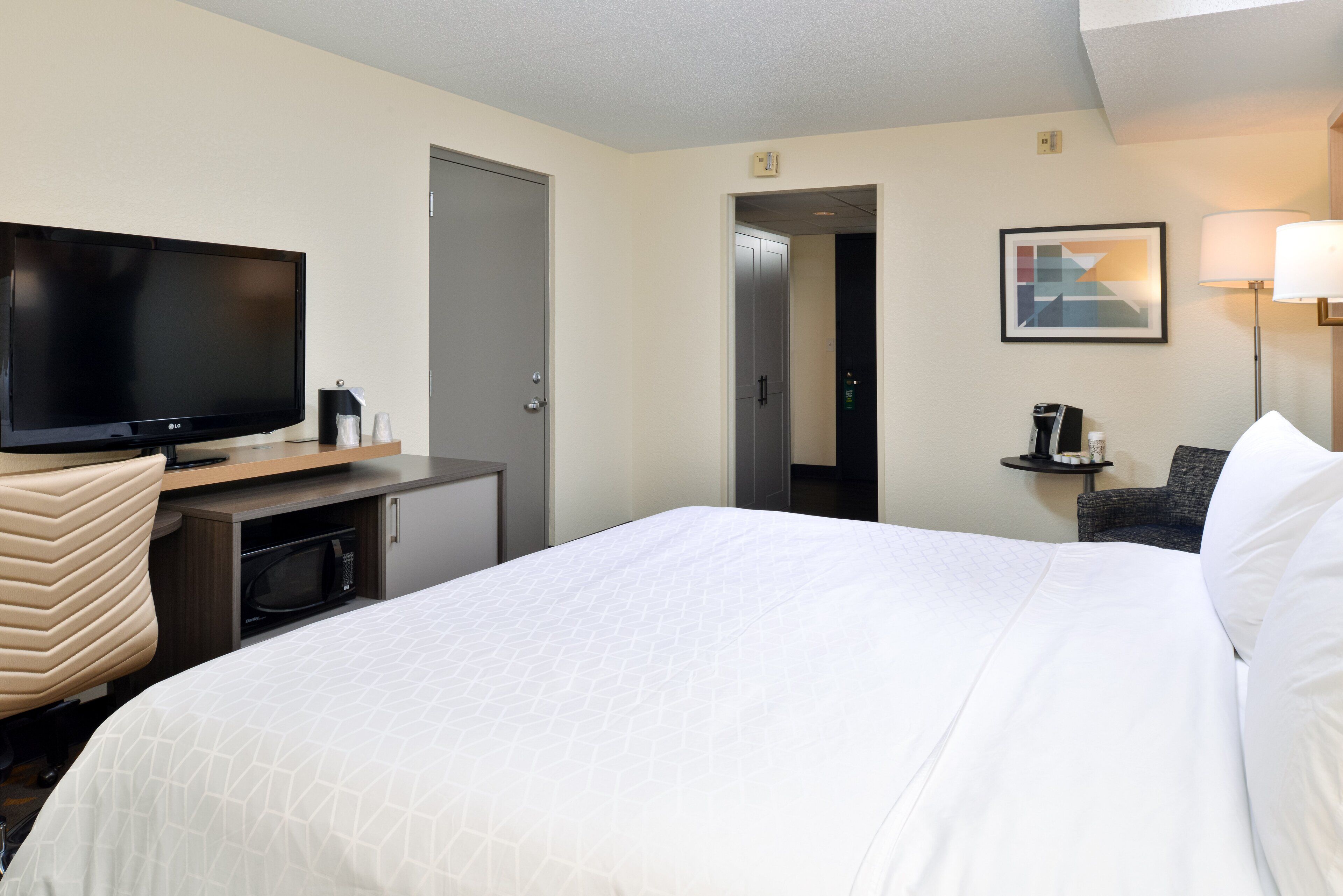 A hotel room with a large bed, a flat-screen TV, a coffee machine, a small table, and a chair.