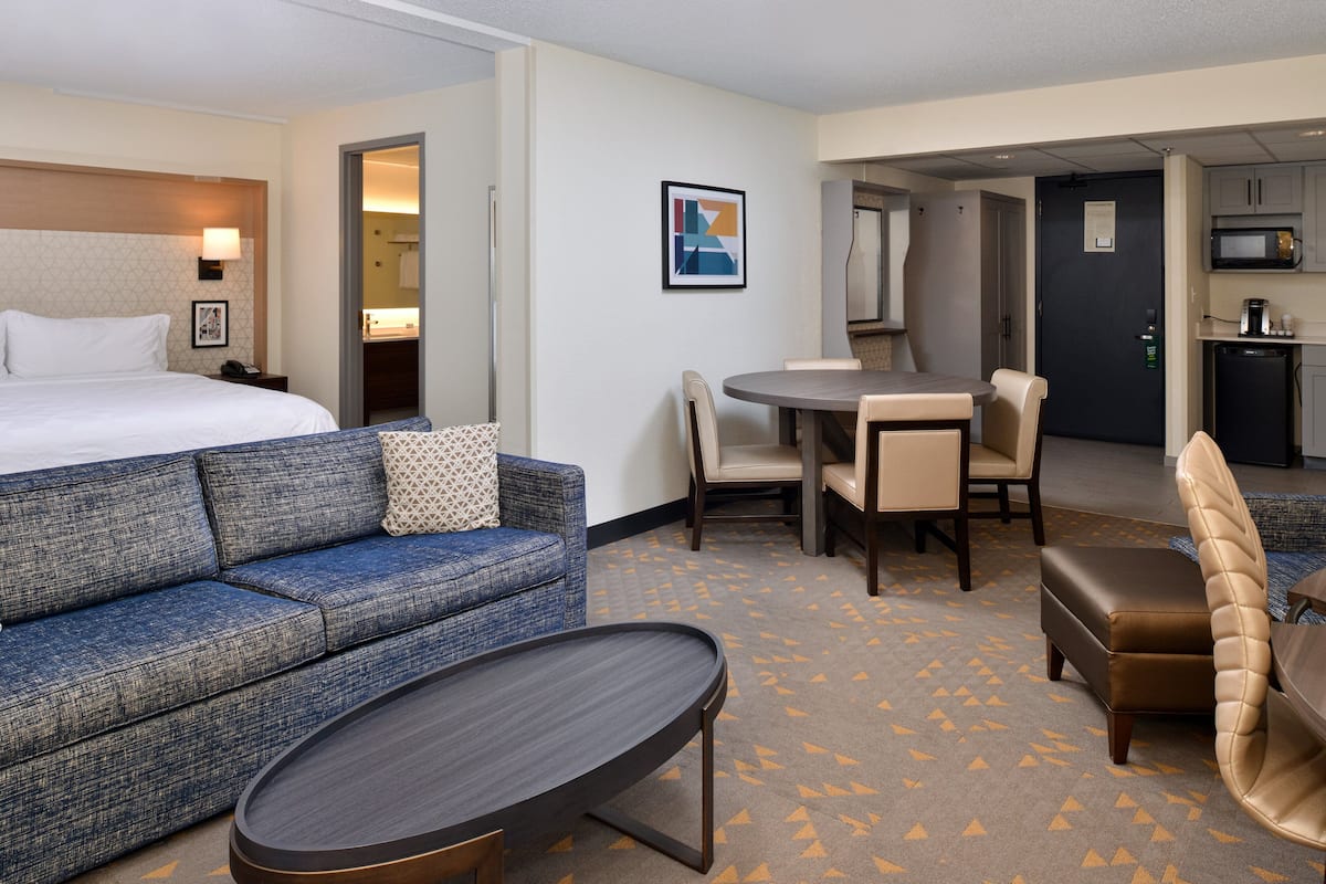 Une chambre d’hôtel comprenant un lit, un canapé, une table à manger avec des chaises, un coin cuisine et une salle de bain visible à travers une porte ouverte.