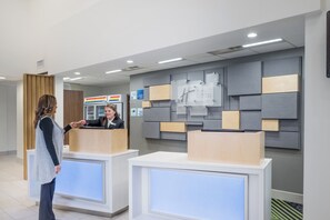 Zona de recepción de hotel con diseño moderno, que incluye un mostrador de recepción, un panel informativo en la pared y un mostrador de registro visible.