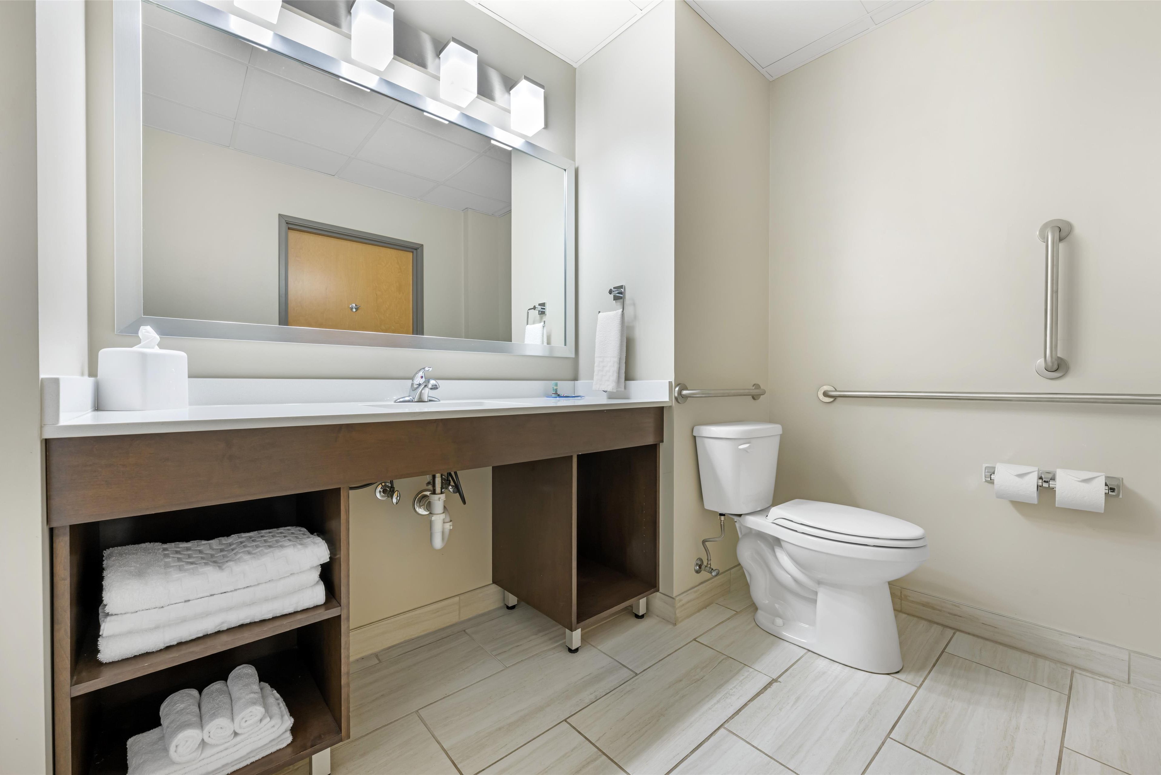 A modern bathroom with a large mirror, white sink, and toilet. There are towels and toiletries on the counter.