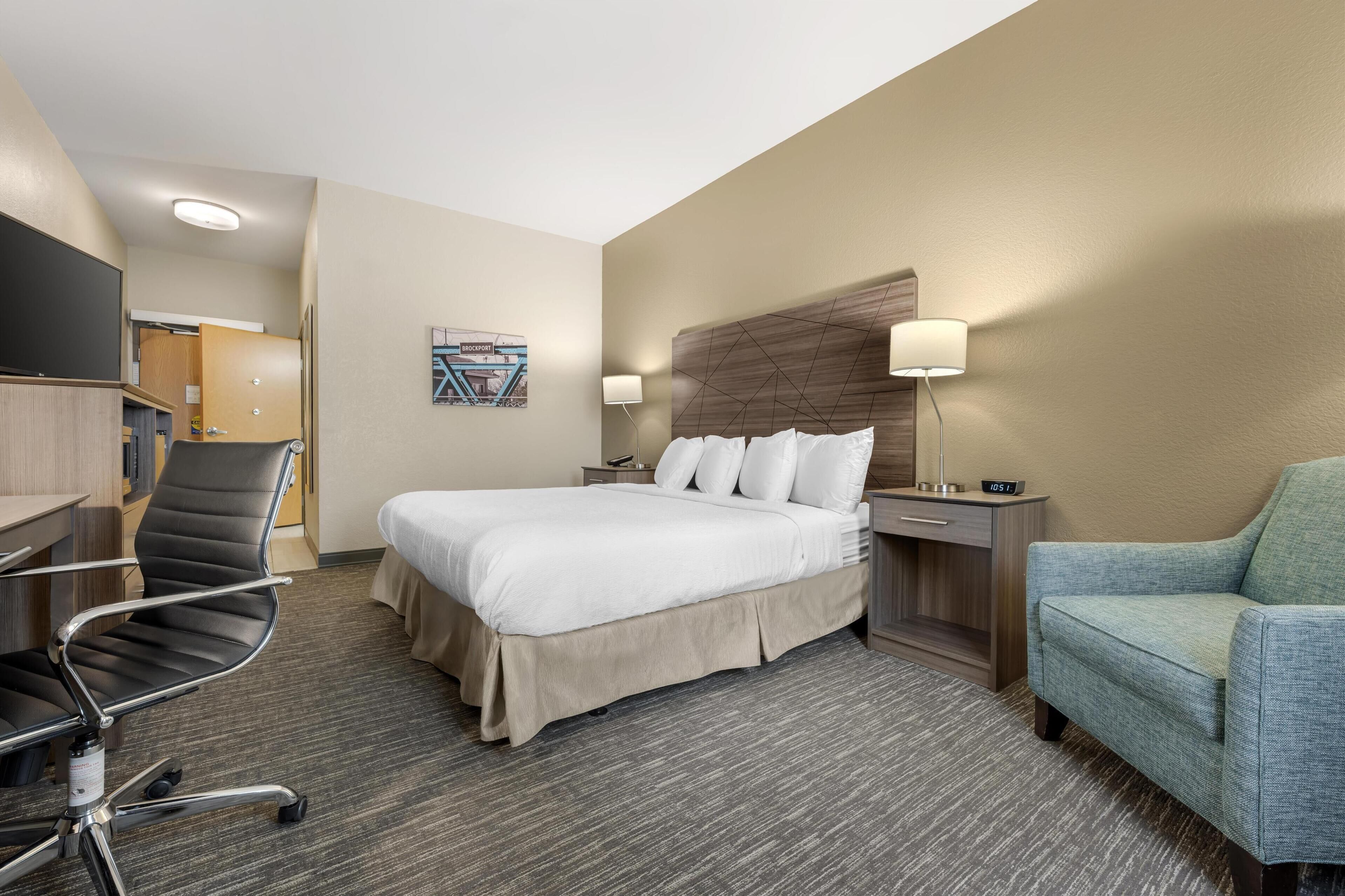 A hotel room with a bed, a desk, a chair, a nightstand, and a lamp.