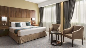 Une chambre d’hôtel dotée d’un grand lit, de deux tables de chevet avec des lampes, d’une table basse ronde, de deux fauteuils et offrant une vue sur l’extérieur grâce à de grandes fenêtres équipées de rideaux.