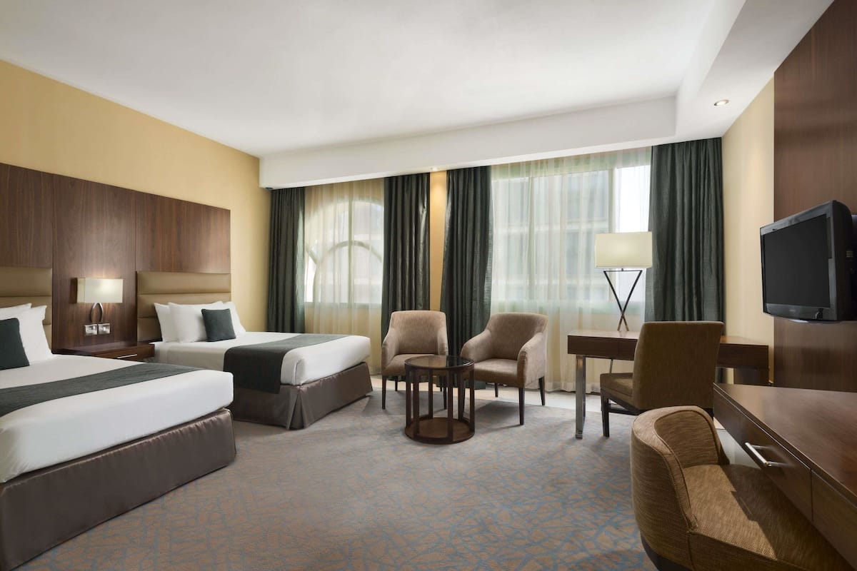 Une chambre d’hôtel avec deux lits, un bureau, deux fauteuils, une télévision et une lampe.
